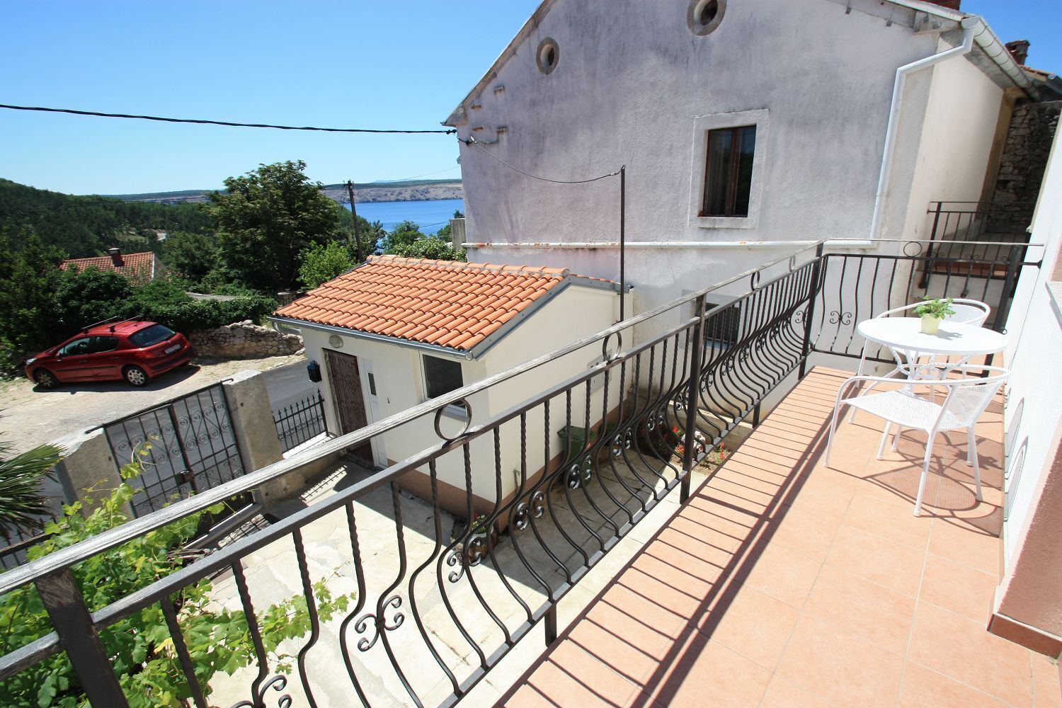 Un balcon avec un sol carrelé, une petite table avec des chaises et une voiture rouge garée à l’extérieur.