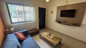 A living room with a sofa, a coffee table, a TV mounted on the wall, and a window with blinds.