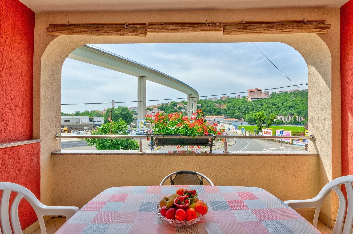 Ein Balkon mit Tisch und Stühlen, einer Obstschale und Blick auf eine Stadtlandschaft mit Grünflächen und einer Brücke.