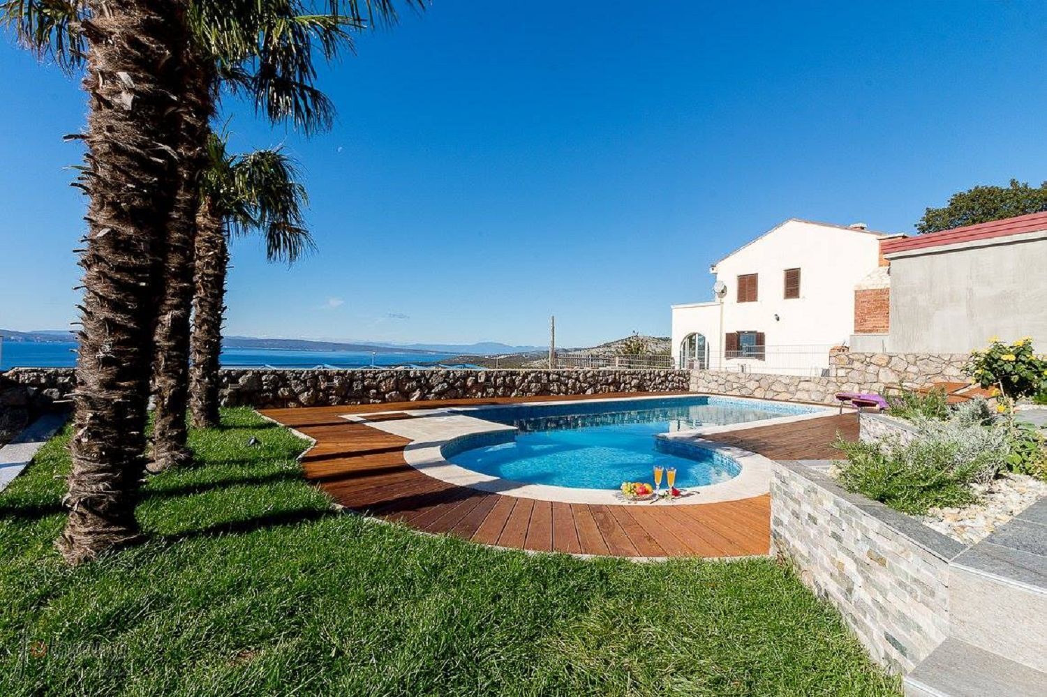 A pool area with a wooden deck, a white building with a red roof, and a view of the sea.