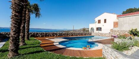 A pool area with a wooden deck, a white building with a red roof, and a view of the sea.