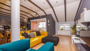 Una sala de estar moderna con un sofá amarillo, sillones azul verdoso y un reposapiés negro. Se observa un área de cocina con una mesa de comedor y sillas al fondo.