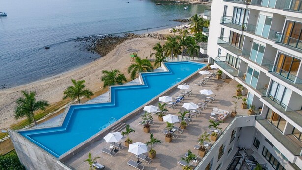 A rooftop pool with a zigzag edge, surrounded by lounge chairs and umbrellas, overlooking a beach and a body of water. - New Studio at la Cruz de Huanacaxtle (Cruz de Huanacaxtle)