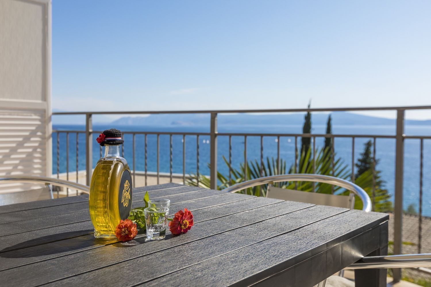 A wooden table with a bottle of olive oil, two glasses, and red flowers on a balcony overlooking the sea.