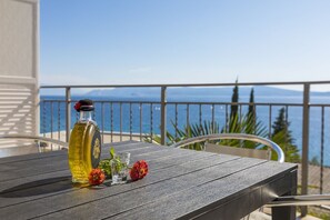 Ein Holztisch mit Olivenöl, zwei Gläsern und roten Blumen auf einem Balkon mit Blick aufs Meer.