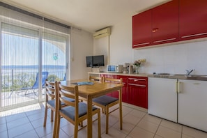 Eine moderne Küche mit roten Schränken, ein Essbereich mit Holzsesseln, einem Fernseher und Blick durch große Fenster.