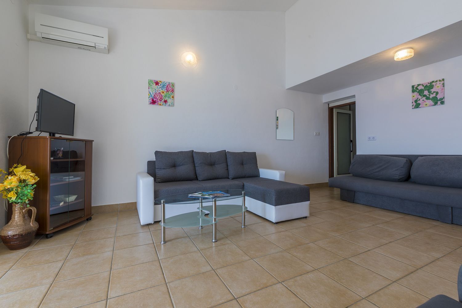 A modern living room with a grey sectional sofa, a glass coffee table, a wooden cabinet with a TV, and a vase with flowers.