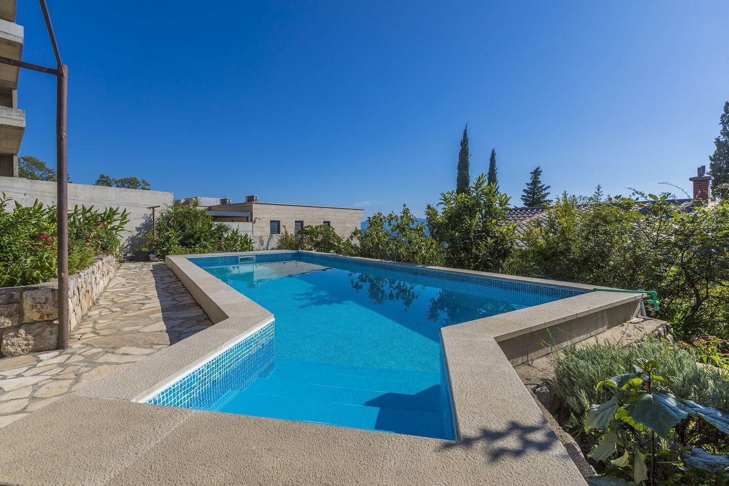 A rectangular swimming pool with clear blue water, surrounded by a stone deck and lush greenery.