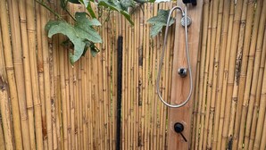 Area doccia esterna recintata da bambù, con doccetta e tubo, una panca in legno e una bottiglia di detergente.