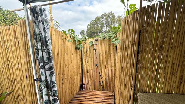 Un'area esterna riparata con un pavimento in legno, una recinzione in bambù, una doccia e un divano in vimini.