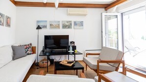 Un salon avec un canapé, un téléviseur, une table basse et un fauteuil. Il y a des tableaux au mur et une fenêtre donnant sur un paysage.