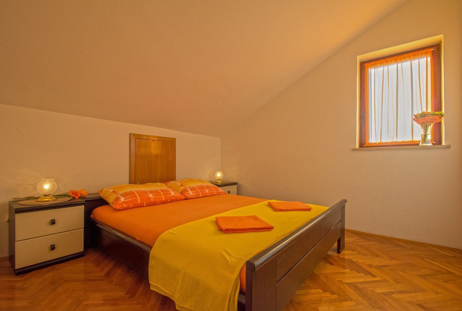 A single bed with an orange bedspread and pillows, a wooden headboard, a nightstand with a lamp, and a window with curtains.