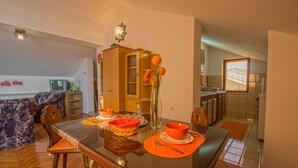 Ein Essbereich mit einem Glastisch, orangefarbenen Tischsets und einer Vase mit Blumen. Im Hintergrund befindet sich ein Holzschrank und ein Fernseher.