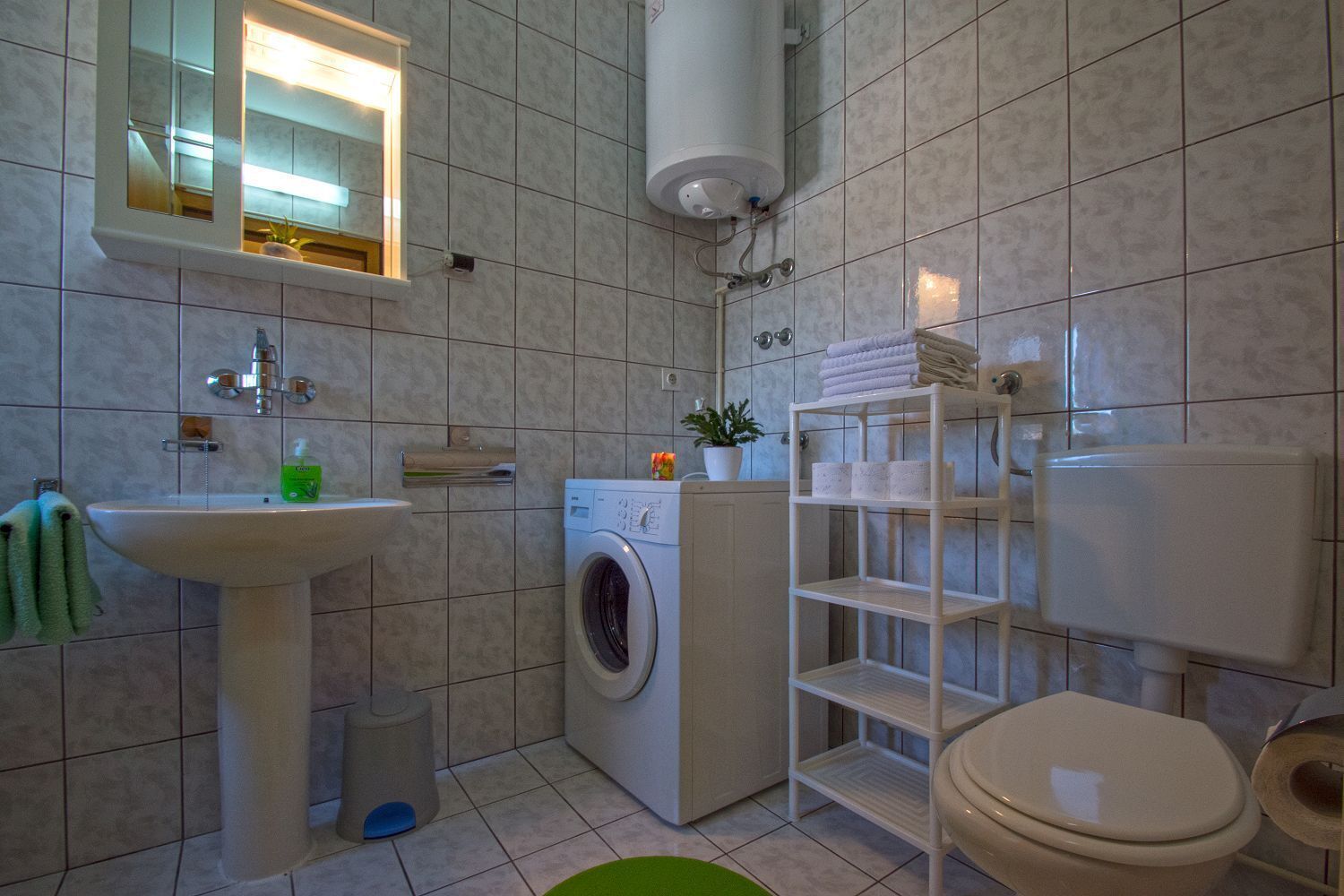 A bathroom with a sink, a washing machine, a toilet, and a shelf with towels.