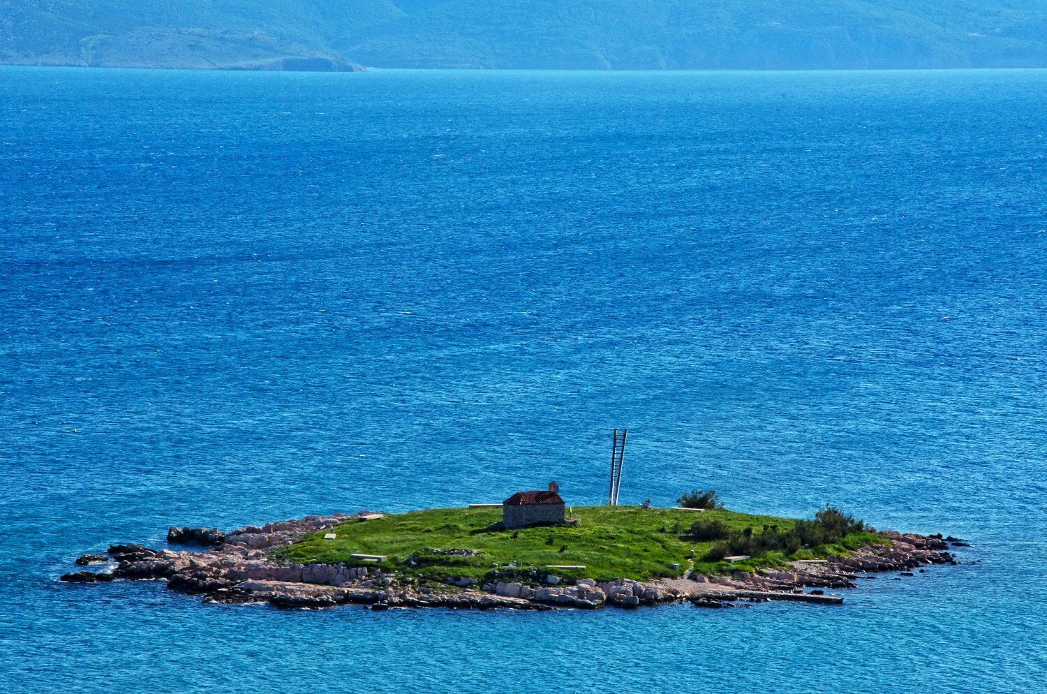 A small island with a single building surrounded by rocky shores and clear blue water.