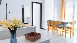 A modern room with a white table, a blue vase with yellow flowers, a woven basket, and a dining area with yellow chairs and a table with a tropical print. - Cozy Flat by the Beach (Jesolo)