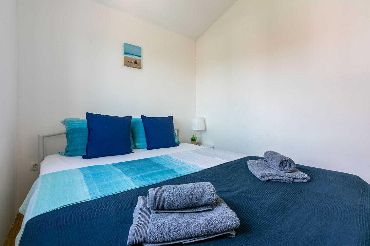 A neatly made bed with blue bedding and pillows, a small wall art, and a lamp on a bedside table.