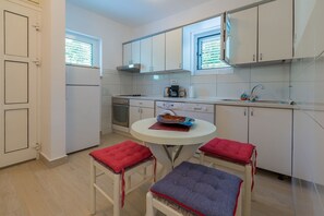 A compact kitchen with white cabinets, a built-in oven, a coffee maker, and a window with blinds. There is a small round table with two stools, each with a red cushion. - Large apartment just 300 metres from the beach with sea views and washing machine (Dramalj)