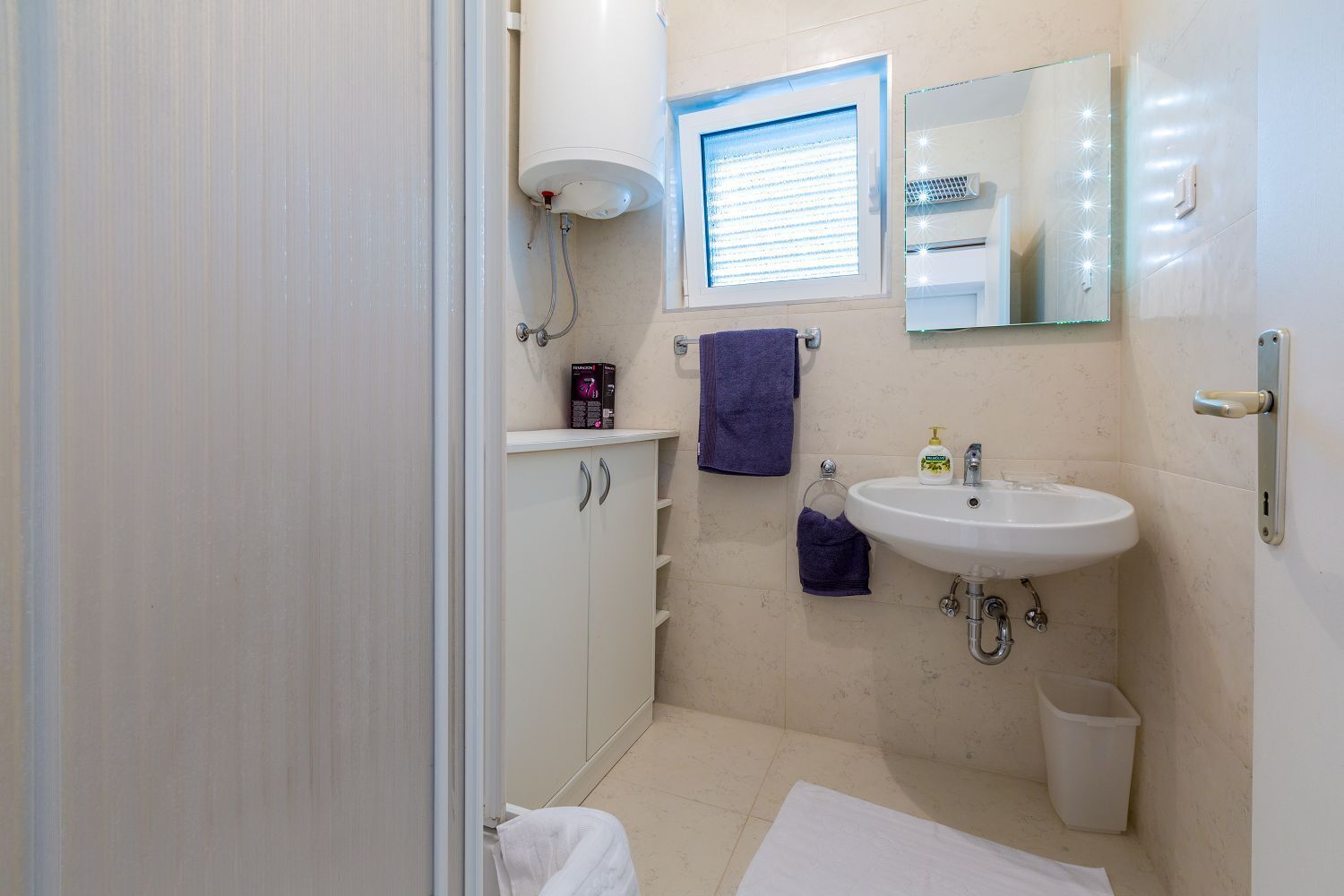 Un baño con un lavamanos blanco, un espejo con iluminación, un perchero para toallas y un armario.