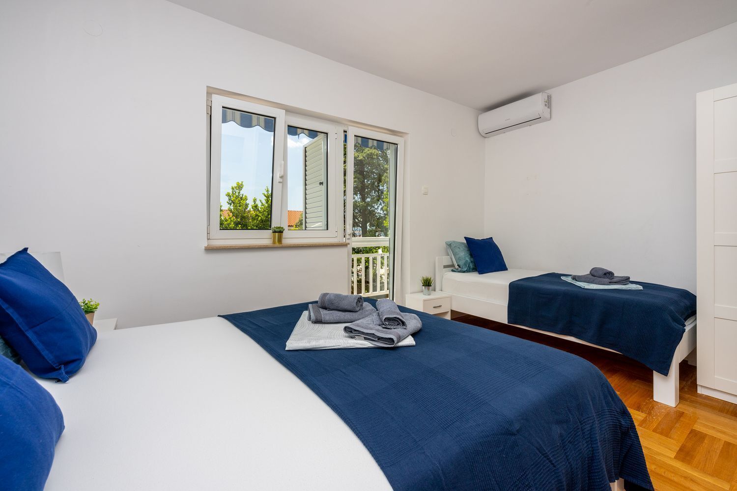 A bedroom with two beds, a window with a view of trees, and an air conditioning unit on the wall.