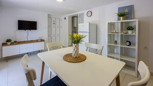 Moderni keittiö, jossa on valkoiset kaapit, sisäänrakennettu uuni sekä valkoinen ruokapöytä tuoleineen. Seinälle on asennettu televisio ja seinällä on myös kello.