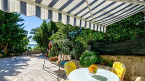 Eine überdachte Terrasse mit Schaukel und einem runden Tisch mit Stühlen, mit Blick auf das Meer.