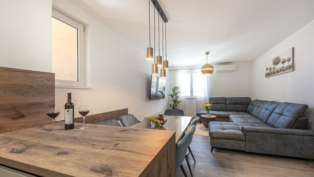 Un espacio de estar moderno con una mesa de comedor de madera, un sofá gris y una zona de cocina con una botella de vino y una copa.