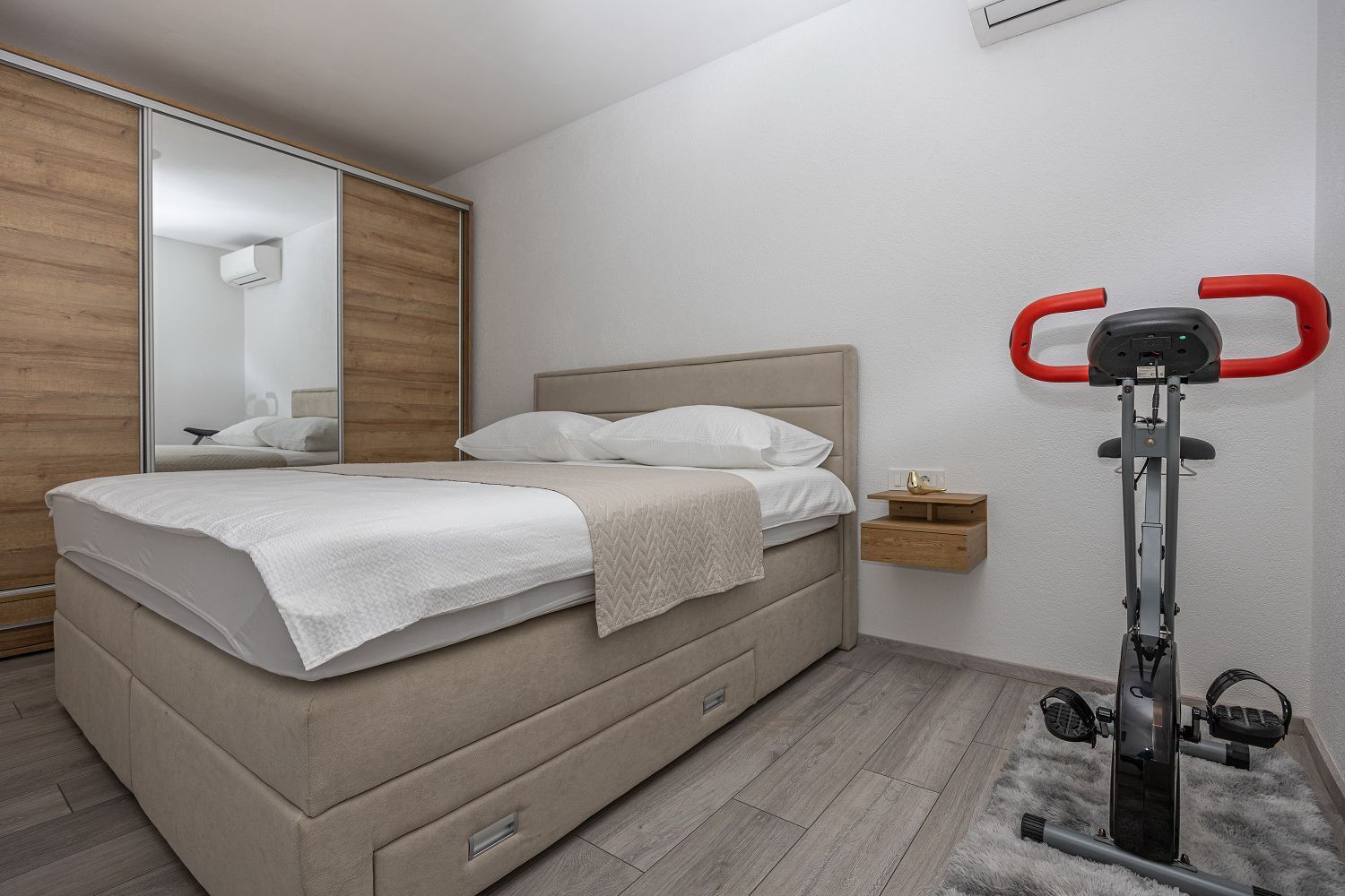 A bedroom with a bed, a nightstand, a mirror, and an exercise bike.