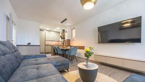 Una sala moderna con un sofá seccional gris, una mesa redonda de centro con un jarrón de flores, un comedor con una mesa de madera y sillas azules, y una cocina al fondo.