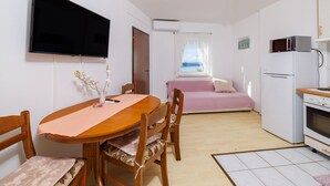 Ein kleines Zimmer mit einem Holzespeisetisch, Stühlen, einem rosa Sofa, einem weißen Kühlschrank, einer Mikrowelle und einem an der Wand befestigten Fernseher.