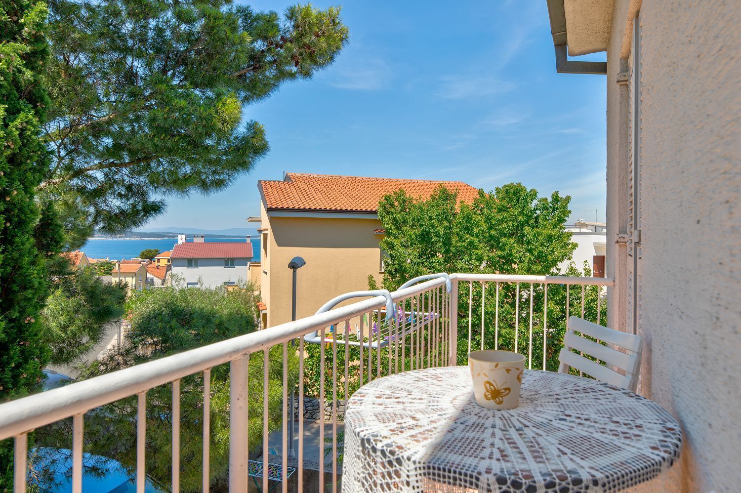 A balcony with a white metal railing, a round table with a lace tablecloth, and a chair. In the background, there is a building with a red roof and a view of the sea.