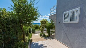 Un étroit sentier mène à un bâtiment à la façade grise, doté d’une fenêtre avec des volets.