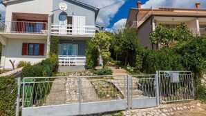 Une maison à deux étages dotée d’un balcon, d’une antenne parabolique et d’un chemin de pierre menant à une entrée fermée par un portail.
