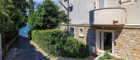 A narrow pathway leading to a stone building with a balcony, surrounded by lush greenery and a clear view of the sea.