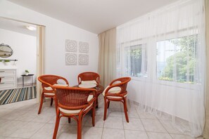 A dining area with wicker chairs and a glass table, a white cabinet with drawers, a small side table with a plant, and a large window with sheer curtains. - Fascinating apartment just 50 metres from the beach with sea view, washing machine, air conditioning, WiFi (Selce)