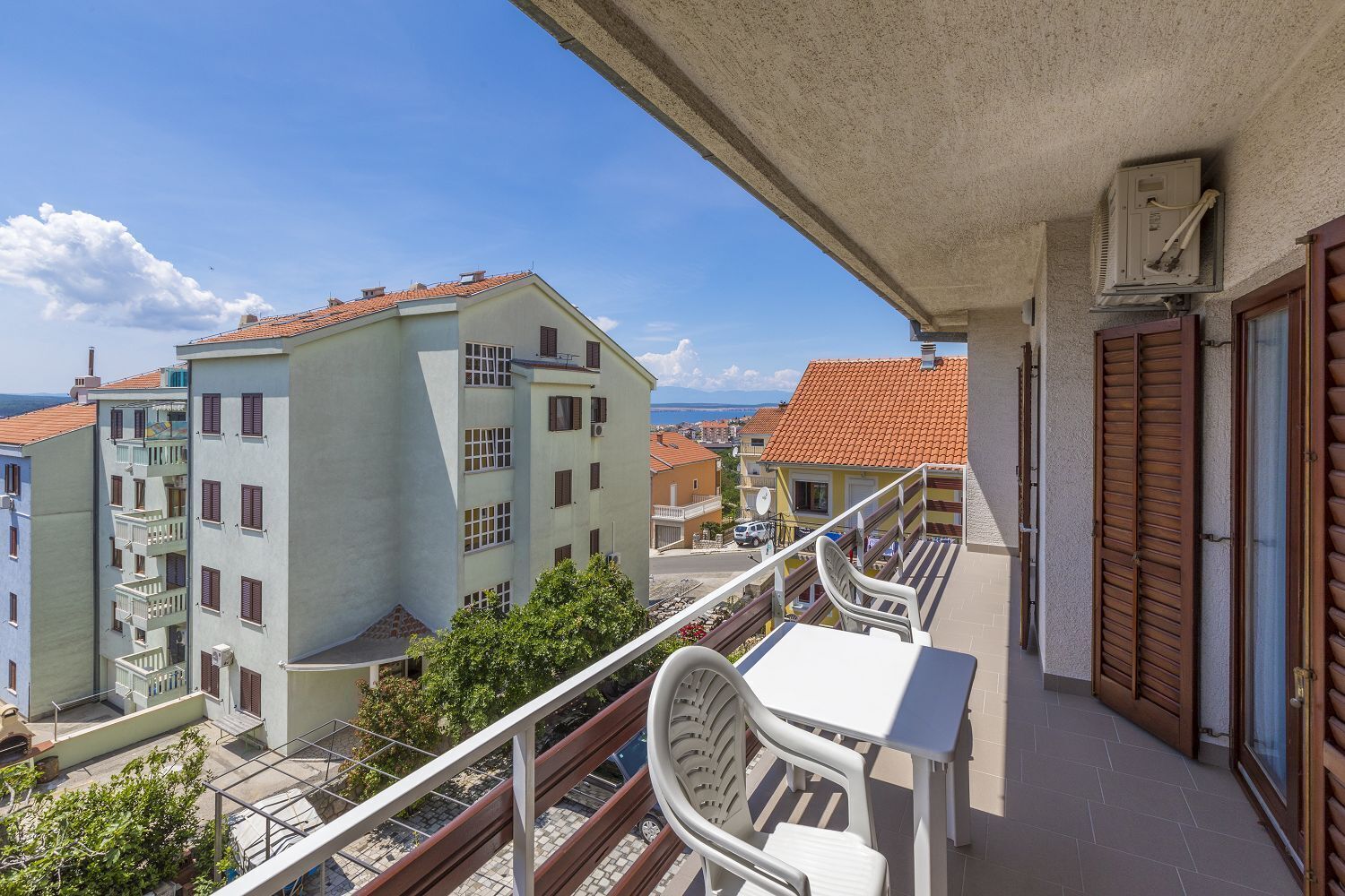 A balcony with a table and chairs, overlooking a coastal town with terracotta-roofed buildings.