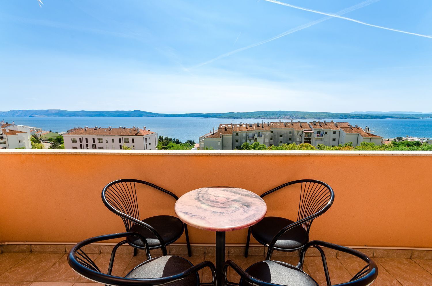 A terrace with a round table and two chairs, overlooking a coastal town with buildings and a clear sky.