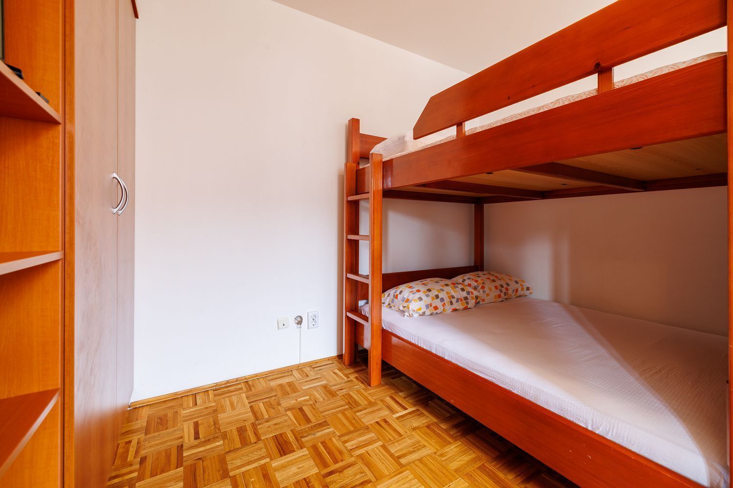 Un lit superposé avec une structure en bois, une petite étagère et un coussin à motifs. On y trouve également une armoire en bois avec une poignée argentée et un parquet à chevrons.
