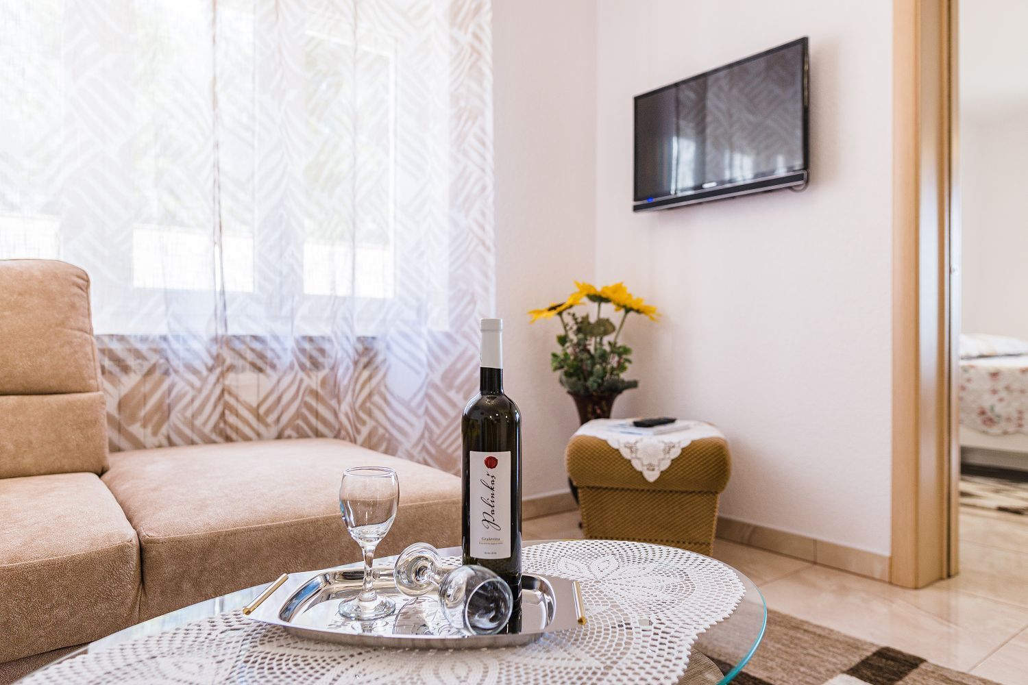 A living room with a sofa, a table with a wine bottle and glasses, a small side table with a plant, and a TV mounted on the wall.