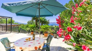 Een terras buiten met een tafel, stoelen, een grote parasol en potplanten.