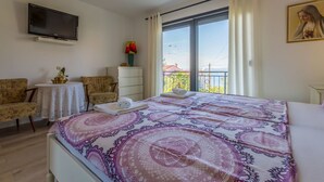 Habitación de hotel con cama, dos sillas, televisor, ventana con cortinas y un cuadro en la pared.