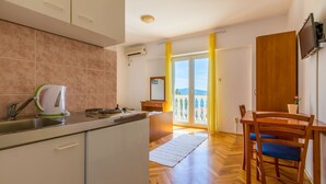 A modern kitchen with a built-in stove, a sink, and a kettle. There is a dining area with a table and chairs. A balcony with yellow curtains is visible through a glass door. - Cosy studio apartment just 300 metres from the beach with sea view and parking space (Crikvenica)