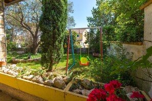 A garden with a swing set, a stone wall, and a yellow-painted building. - Cute studio apartment just 300 metres from the beach with a romantic balcony (Crikvenica)