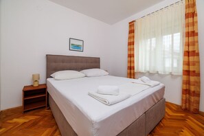 A neatly made bed with white linens and a folded towel, a bedside table with a lamp, and a window with striped curtains. - Cosy apartment just 300 metres from the beach with sea views and a children's swing (Crikvenica)
