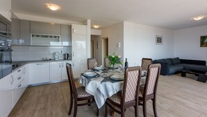 Een moderne keuken met witte kasten, een eetgedeelte met een tafel voor vier personen, en een donkergrijze bank in de woonkamer.