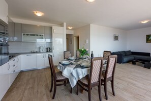 Eine moderne Küche mit weißen Schränken, ein Essbereich mit Tisch für vier Personen und ein dunkelgraues Sofa im Wohnzimmer.