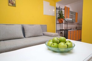 Ein Wohnzimmer mit grauem Sofa, einem weißen Tisch mit einer Schüssel grüner Äpfel und einer Küche im Hintergrund mit orangefarbenen Schränken.