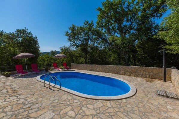 A swimming pool with stone flooring, surrounded by red lounge chairs and umbrellas, with trees in the background.