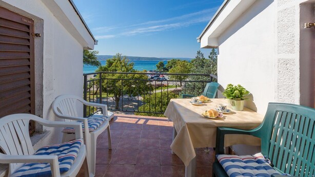 Ein Balkon mit weißen Stühlen, einem Tisch für zwei Personen und Blick auf das Meer und grüne Vegetation.
