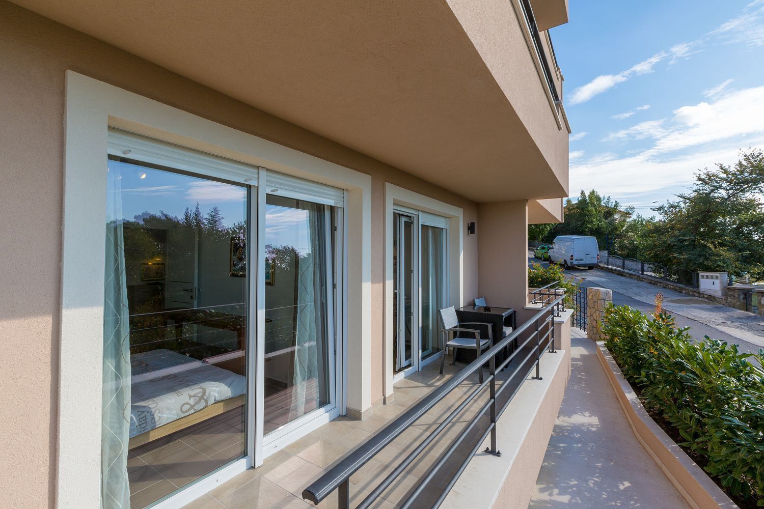 A balcony with a glass door, a small table with chairs, and a view of a street with parked cars.